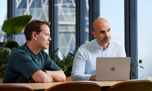 Ahmed, avec son MacBook devant lui, assis à une table avec quelqu’un et regardant quelqu’un d’autre hors champ.