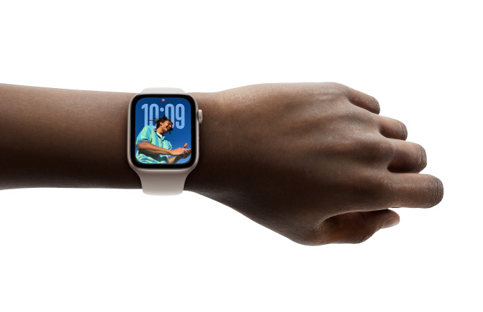 Nahaufnahme einer Apple Watch in Roségold mit Gummiarmband am Handgelenk einer Person, das Display zeigt die Uhr App im neuen Liquid Glass Design