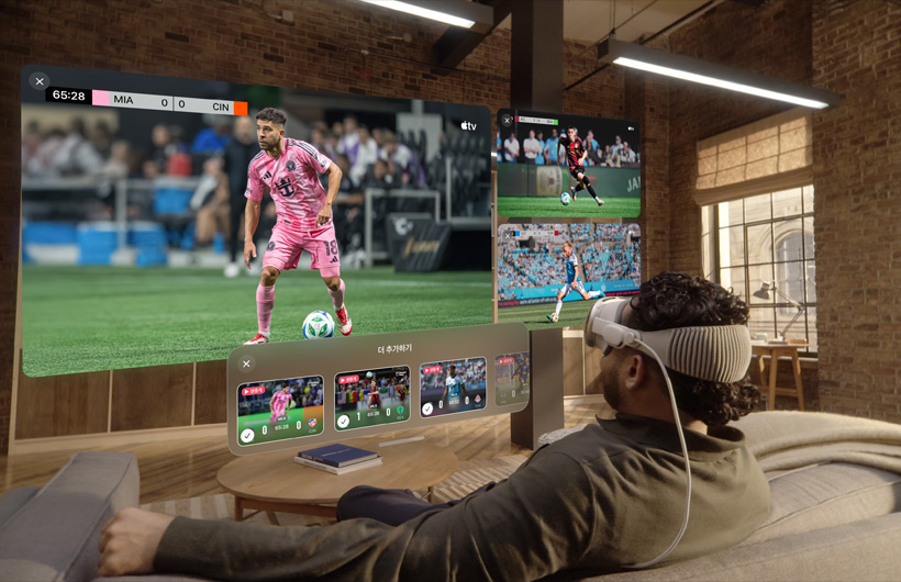 Apple Vision Pro를 착용한 사람이 거실 소파에 앉아 있는 모습. 그 앞에는 큰 동영상 화면과 옆으로 두 개의 작은 동영상 화면이 나란히 떠 있으며, 각각 다른 스포츠 경기를 보여줍니다. 큰 화면 밑에는 라고 적힌 선택 창이 있습니다. 더 추가하기