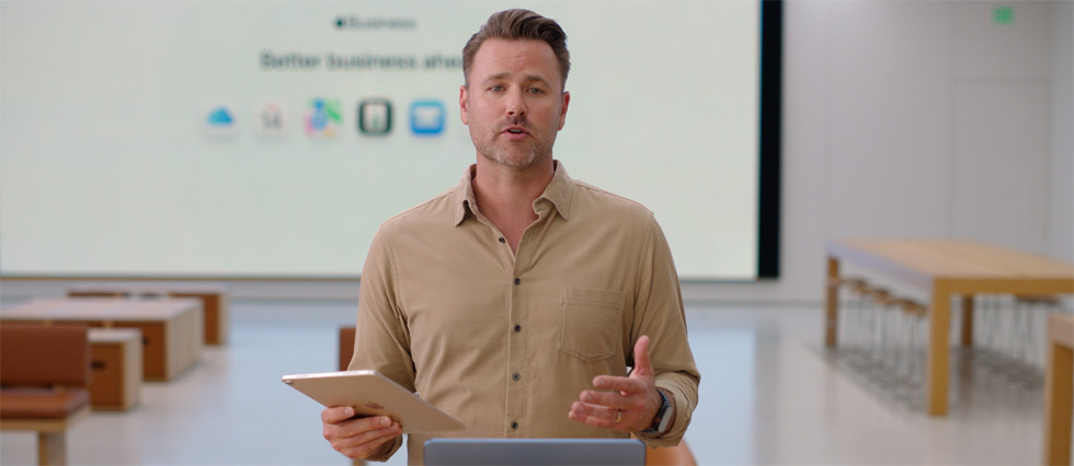 Presenter speaks while referencing an iPad and a large presentation screen displays various business app icons.