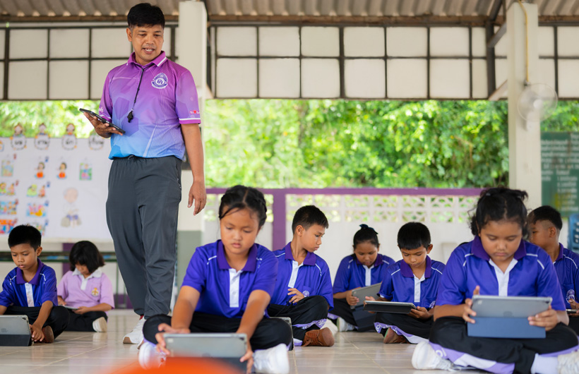ในห้องเรียนกลางแจ้ง ครูพลศึกษาเดินดูนักเรียนที่นั่งอยู่บนพื้นและใช้งาน iPad