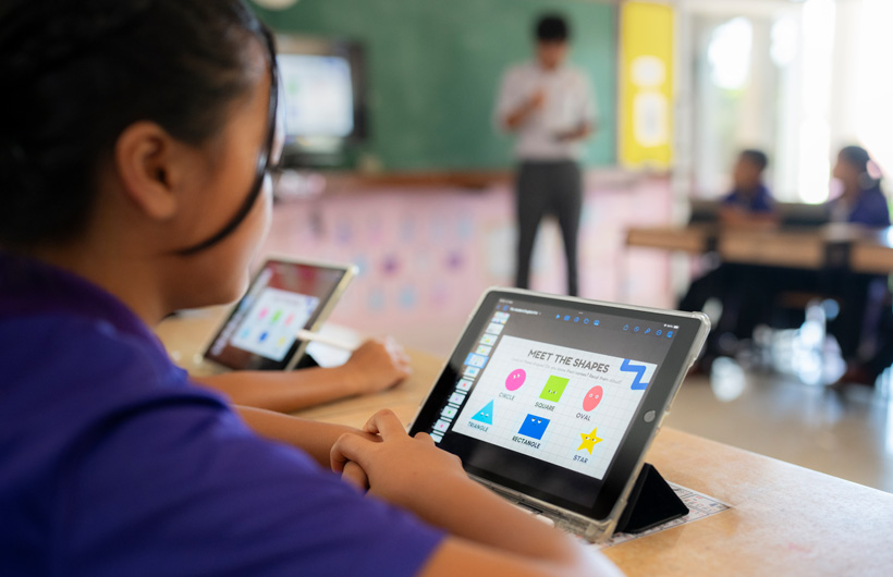 ภาพระยะใกล้แสดงนักเรียนนั่งอยู่ที่โต๊ะเรียนขณะกำลังใช้ iPad โดยบนหน้าจอมีรูปทรงหลากสีสัน พร้อมชื่อเรื่อง 
