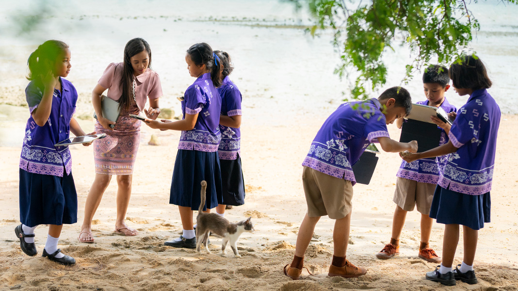 นักเรียนชั้นประถมศึกษาจำนวน 6 คนในชุดนักเรียนสีม่วงสดยืนถือ iPad อยู่บนหาดทราย โดยผู้สอนกำลังแนะนำนักเรียนคนหนึ่งบน iPad และมีแมวเดินผ่านพวกเขา