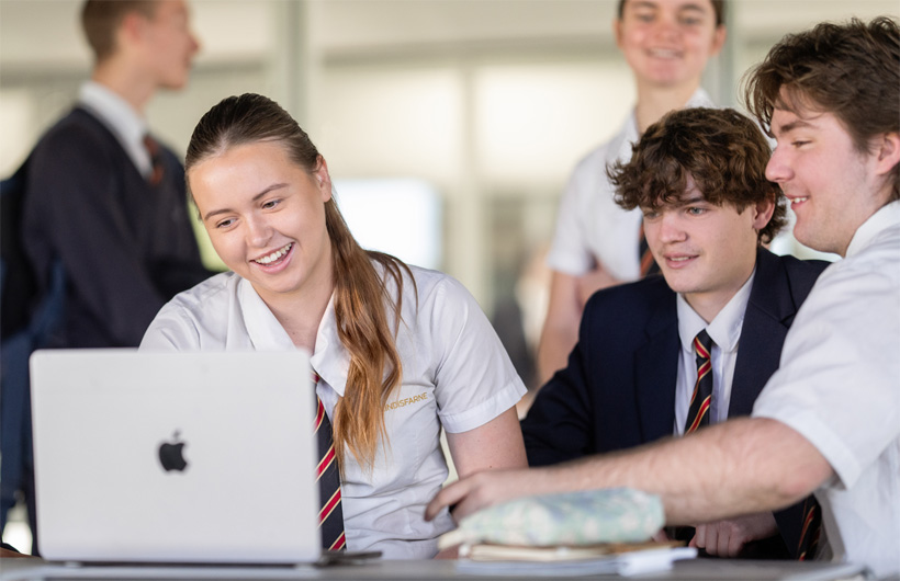 กลุ่มนักเรียนในชุดเครื่องแบบของโรงเรียนรวมตัวกันรอบ MacBook ด้วยสีหน้ายิ้มแย้มและกำลังช่วยกันทำงาน